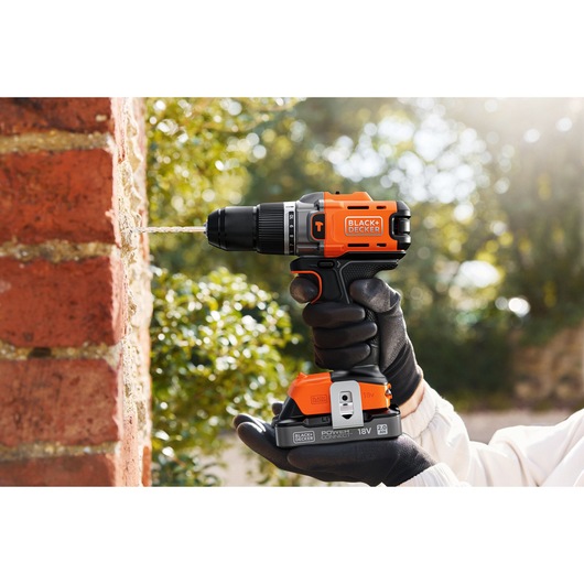 BLACK+DECKER cordless drill used on a brick wall, held by a gloved hand outdoors.