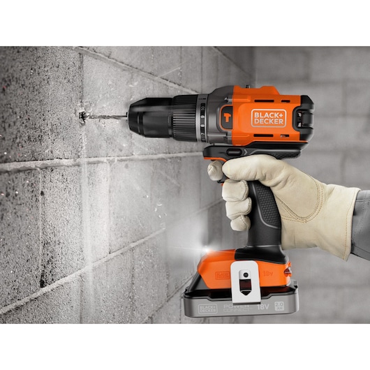 Cordless BLACK+DECKER drill in use, drilling into a concrete wall, held by a gloved hand.