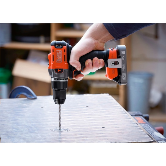 Hand using BLACK+DECKER cordless drill to make a hole in a metal sheet in a workshop.