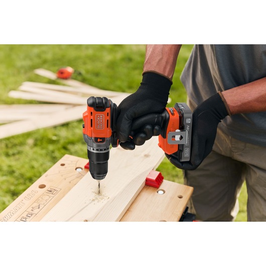 Person drills wood outdoors with a BLACK+DECKER cordless drill, wearing black gloves for safety.