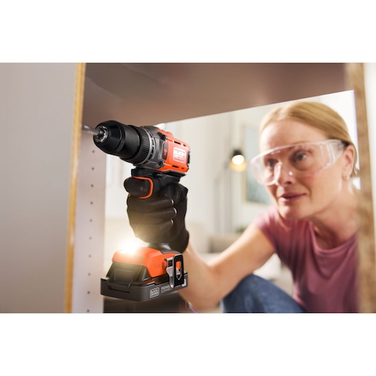Person using a BLACK+DECKER cordless drill to assemble furniture, wearing gloves and safety glasses.