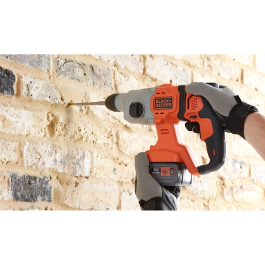 Photo of BLACK+DECKER BCD900M1K cordless hammer drill in use on brick wall, orange and black design, 18V battery visible.