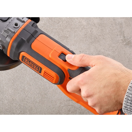 Photo of a hand operating the power switch on a BLACK+DECKER angle grinder BCG720M1 against a gray background.