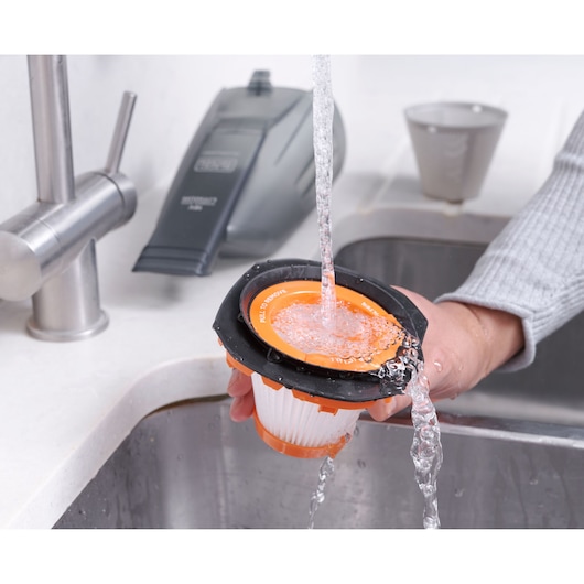 BLACKDECKER BCHV001C1 vacuum filter being rinsed under running water in a kitchen sink, vacuum in background.