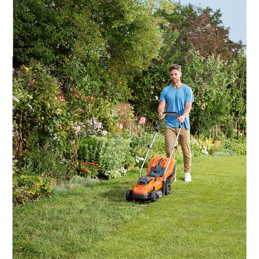 Person mowing lawn with BLACKDECKER BCMW33182L2 orange cordless lawn mower in a garden.