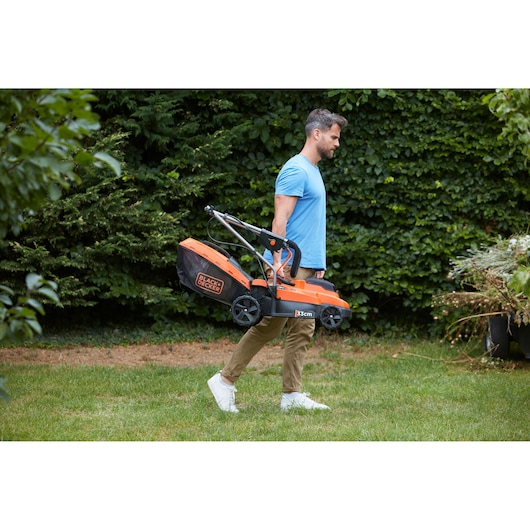 Person carrying BLACK+DECKER BCMW33182L2 orange and black lawn mower with folding handle in a garden.