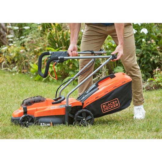 BLACK+DECKER BCMW33182L2 orange cordless lawn mower with black grass collection bag, shown in use on grass.