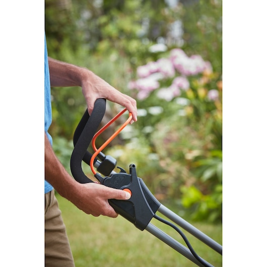 Close-up photo of BLACKDECKER BCMW33182L2 lawn mower handle with black and orange controls held by a person in a garden.