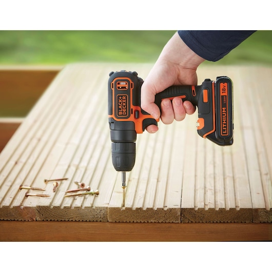 Photo of BLACK+DECKER BDCD18 cordless drill in use on wooden boards, visible screws beside, outdoor setting.