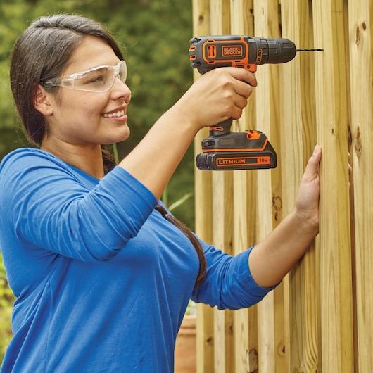 Person using BLACK+DECKER BDCDC18B cordless drill to drill into outdoor wooden fence.