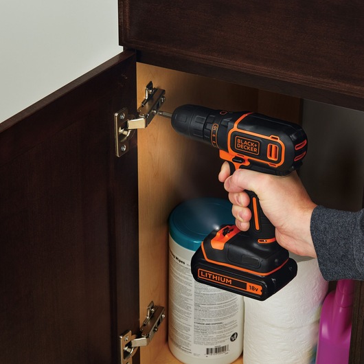 BLACK+DECKER BDCDC18K1B cordless drill used to install cabinet hinge, black and orange design, close-up view.