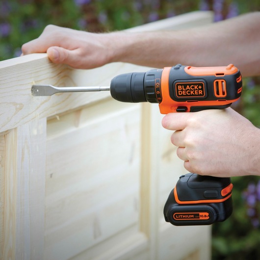 BLACKDECKER BDCDD12B cordless drill in use drilling into light wood outdoors, featuring orange and black design.