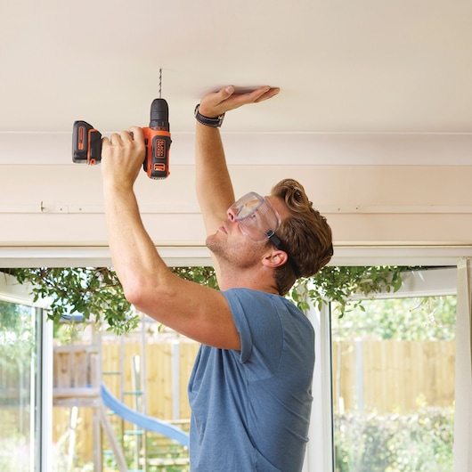 BLACKDECKER BDCDD12B cordless drill used on ceiling indoors by person in blue shirt.