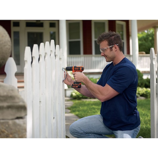 BLACKDECKER BDCDD12KB cordless drill used to fix metal hinge on white wooden fence outdoors.