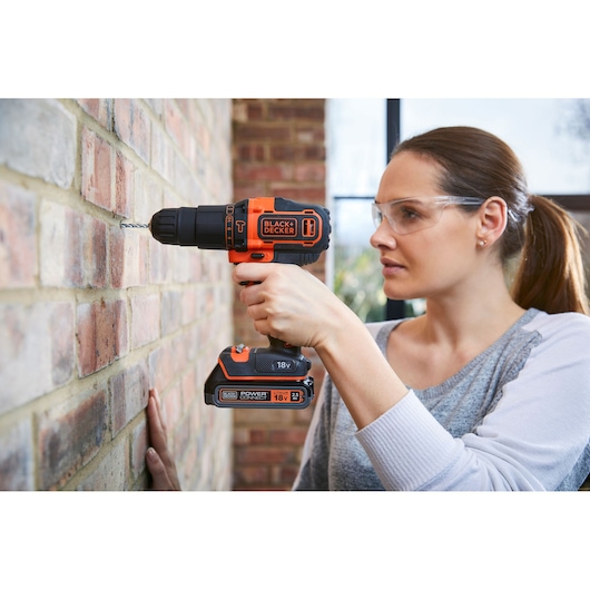Person using BLACK+DECKER BDCHD18K1B2 cordless hammer drill on brick wall indoors.