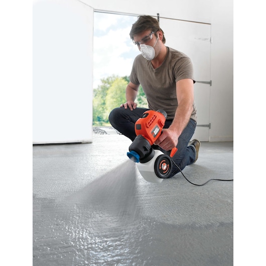 Person using BLACKDECKER BDPS200 electric paint sprayer on indoor floor, orange and black design, spraying gray coating.