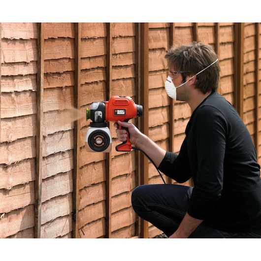 BLACK+DECKER BDPS400 paint sprayer in use on wooden fence, held by person wearing mask.