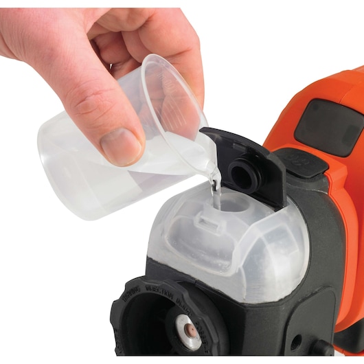 A hand is pouring liquid from a clear plastic cup into the transparent reservoir of an orange and black BLACK+DECKER device.