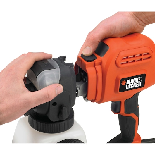 Close-up of two hands assembling a BLACK+DECKER orange spray gun, attaching the nozzle to the main body, with a white container at the bottom.