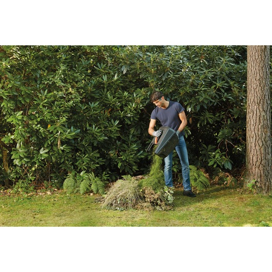 BLACKDECKER BEMW351GL2 lawnmower grass box being emptied by a man onto a garden compost pile outdoors.