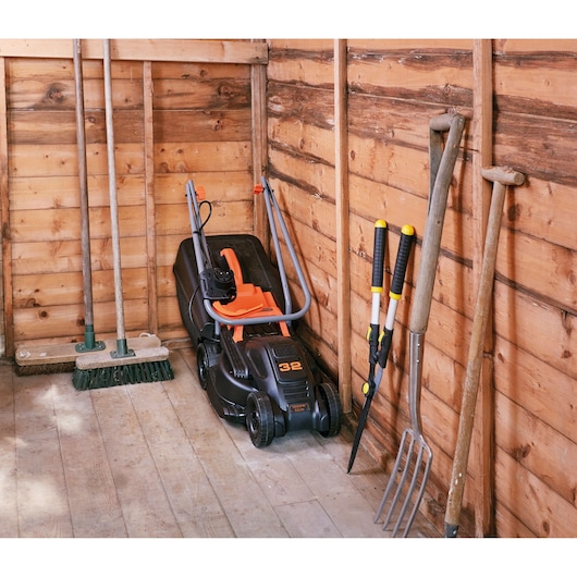 BLACKDECKER BEMW351GL2 lawn mower with folded handle stored in a wooden shed, surrounded by garden tools.