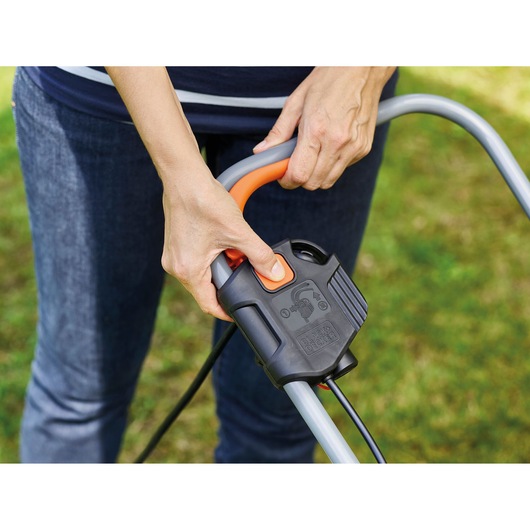 Close-up photo of a person pressing the start button on BLACK+DECKER BEMW351GL2 lawn mower handle outdoors.