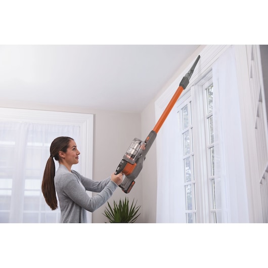 BLACKDECKER BHFEV182C cordless stick vacuum with long orange nozzle cleaning window frame indoors.