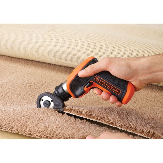 BLACK+DECKER CSCA3 electric rotary cutter trimming beige carpet, close-up of hand holding tool.