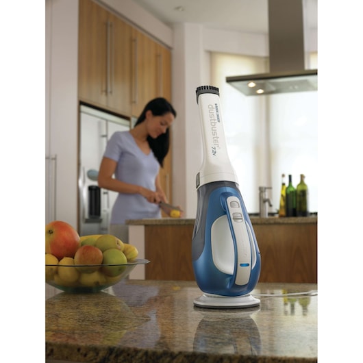 BLACKDECKER DV7210N handheld vacuum cleaner standing on a kitchen countertop with fruit bowl and woman in background.
