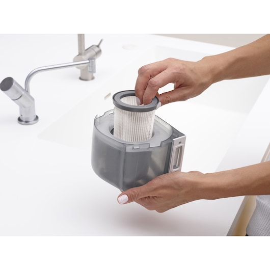 Photo of hands removing filter from BLACKDECKER DVJ325BFS vacuum dust container near a kitchen sink.