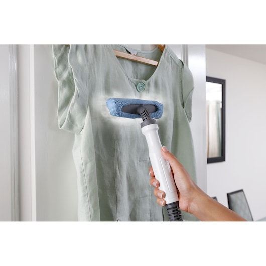 A hand holding a steam cleaning device with a blue cloth attachment, using it to steam clean or refresh a light green shirt hanging on a wooden hanger in a home setting.