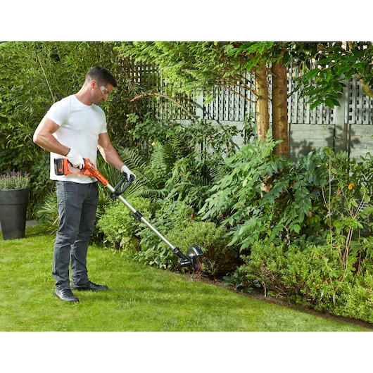 BLACKDECKER GLC3630LB cordless grass trimmer in use on garden lawn near shrubs and bushes.