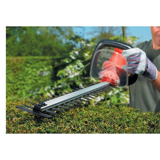 Close-up of a person trimming a hedge with a BLACK+DECKER electric hedge trimmer outdoors. The person is wearing gloves and using the tool to cut the dense green foliage.