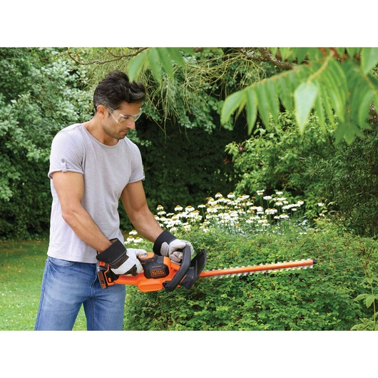 Photo of person using BLACK+DECKER GTC18452PC hedge trimmer outdoors, trimming green bushes in a garden.