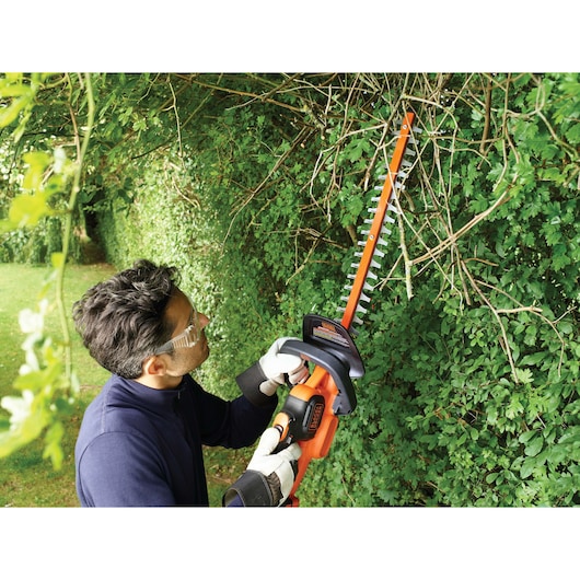 Person using BLACK+DECKER GTC18452PC cordless hedge trimmer on tall green hedge outdoors.