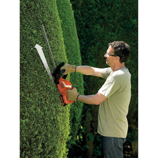 A person wearing gloves using a BLACK+DECKER cordless hedge trimmer to trim a tall, dense hedge outdoors. The hedge trimmer is orange and black, and an arrow indicates the direction of cutting.