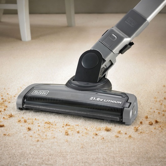 A close-up of a BLACK+DECKER cordless vacuum cleaner with a 21.6V lithium battery, cleaning crumbs from a beige carpet.