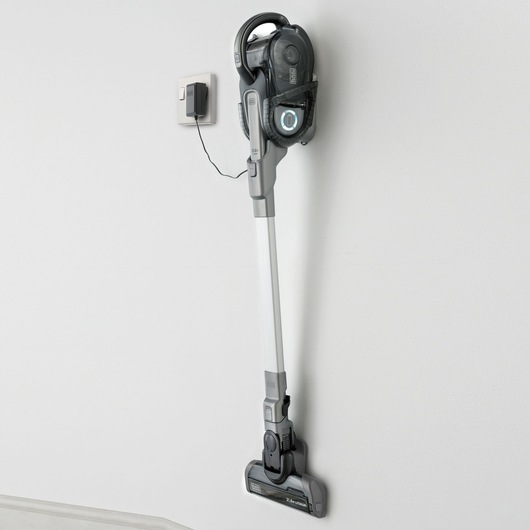 A BLACK+DECKER cordless stick vacuum cleaner mounted vertically on a white wall and plugged in for charging.