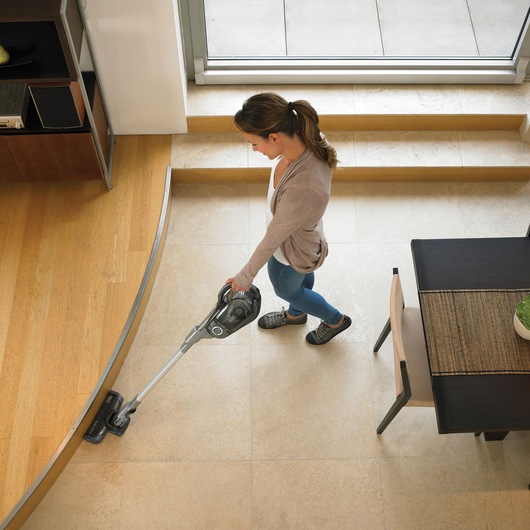 A person vacuuming a room with a BLACK+DECKER cordless stick vacuum cleaner (model HVFE2150LB), transitioning from a tiled floor to a wood floor in a modern home interior.