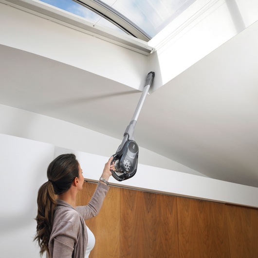 A person using a BLACK+DECKER cordless vacuum cleaner with an extended attachment to clean the upper corners of a room near a window.