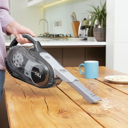 A hand using a BLACK+DECKER cordless handheld vacuum cleaner to pick up crumbs from a wooden kitchen table. There is a blue mug and a plate with food in the background.