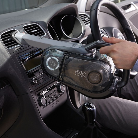 A person is using a BLACK+DECKER handheld vacuum cleaner with a long nozzle attachment to clean the air vent inside a car.
