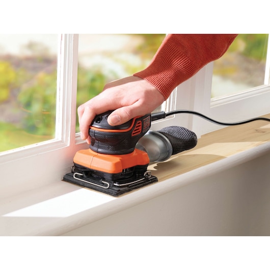 Photo of BLACK+DECKER KA450 sheet sander in use on a wooden windowsill, showing ergonomic grip and dust bag.