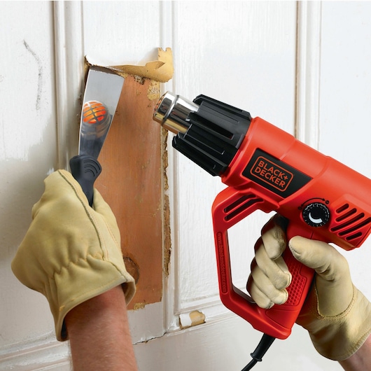 BLACK+DECKER KX2001K heat gun used to strip paint from wood surface, gloved hands holding tool and scraper.