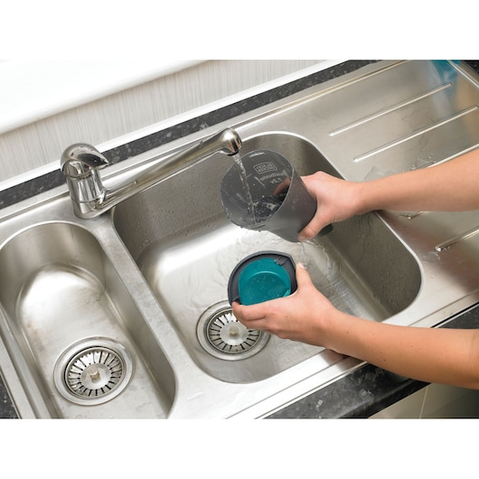 Photo of BLACK+DECKER NV7210N vacuum filter and dust container being washed under a faucet in a stainless steel sink.
