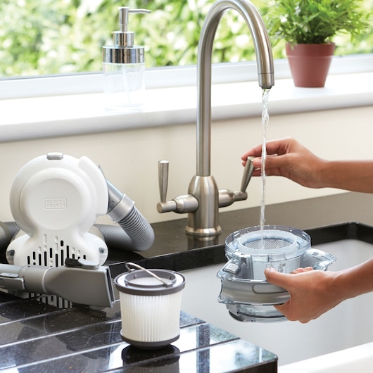 Photo of BLACKDECKER PD1020L handheld vacuum filter and canister being cleaned under faucet, parts on black countertop.