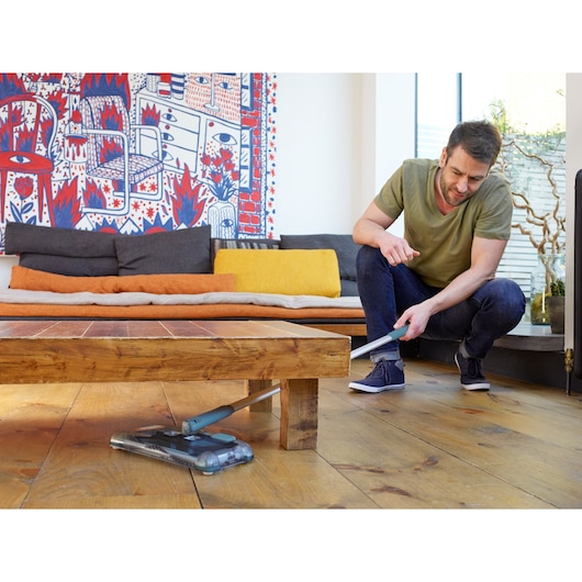BLACKDECKER PSA115B floor sweeper cleaning under wooden coffee table in living room.