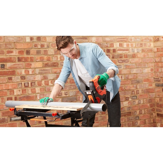 Photo of a person using BLACK+DECKER RS890K electric saw to cut wood on a workbench, brick wall background.