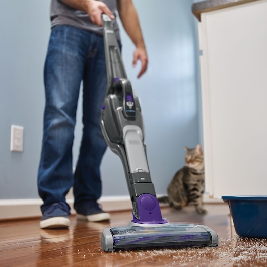 BLACKDECKER SVJ520BFSP stick vacuum cleaning pet litter from hardwood floor, person in background, indoor setting.