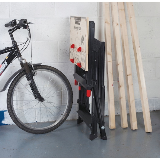 BLACKDECKER WM1000 folding workbench with wood top, black frame, and red clamps, stored upright against a wall.